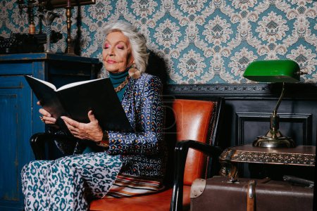 Téléchargez les photos : Femme âgée élégante dans le livre de lecture d'usure de luxe tout en se relaxant à la maison de style rétro - en image libre de droit