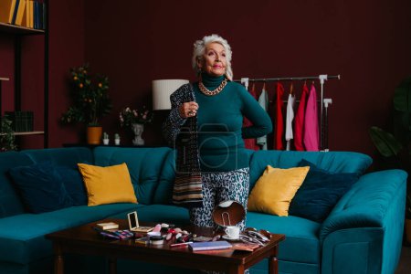 Foto de Mujer mayor de confianza con maquillaje que lleva ropa de moda mientras está de pie en la sala de estar moderna - Imagen libre de derechos