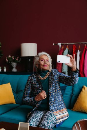 Téléchargez les photos : Femme âgée à la mode faisant selfie par téléphone intelligent tout en étant assis sur le canapé au salon moderne - en image libre de droit