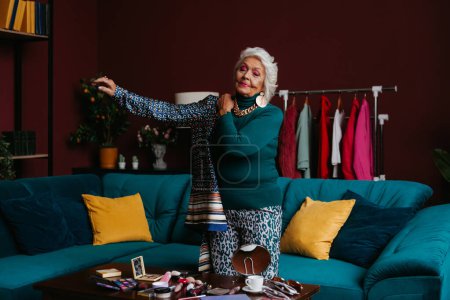 Téléchargez les photos : Femme âgée à la mode avec maquillage portant des vêtements à la mode tout en se tenant debout dans le salon moderne - en image libre de droit