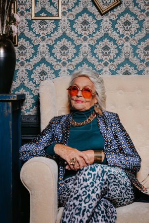 Téléchargez les photos : Femme âgée élégante en tenue tendance et de luxe assise sur un canapé confortable à l'intérieur de la maison de style rétro - en image libre de droit