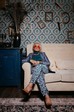 Téléchargez les photos : Femme âgée à la mode en tenue tendance et de luxe assise sur un canapé confortable dans une maison de style rétro - en image libre de droit