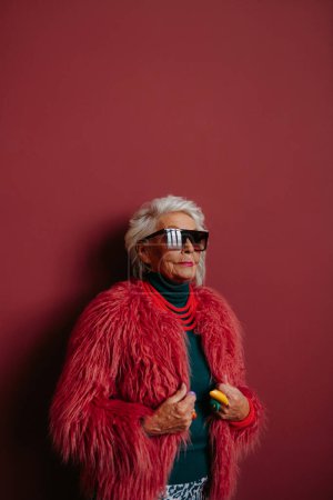 Téléchargez les photos : Femme âgée à la mode avec des lunettes élégantes et des vêtements tendance debout sur fond rouge - en image libre de droit