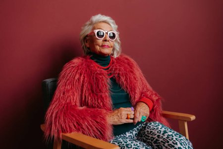 Photo for Confident senior woman in fashionable wear looking cool and trendy while sitting against red background - Royalty Free Image