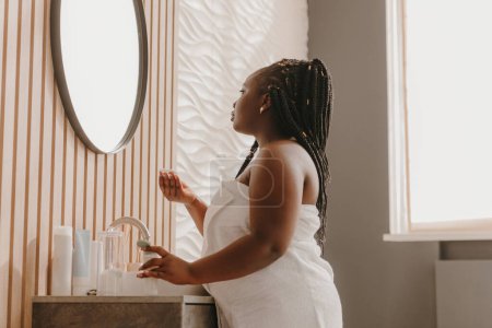 Foto de Mujer africana con curvas de confianza disfrutando de los procedimientos de rutina de la mañana y mirando al espejo en el baño - Imagen libre de derechos