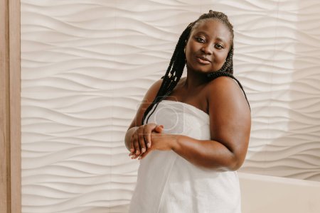 Téléchargez les photos : Confiant dodu plus la taille femme africaine couverte de serviette appliquer de la crème hydratante sur place dans la salle de bain - en image libre de droit