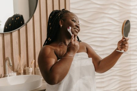 Téléchargez les photos : Confiant plus la taille femme africaine couverte de serviette massant visage avec rouleau à la salle de bain domestique - en image libre de droit