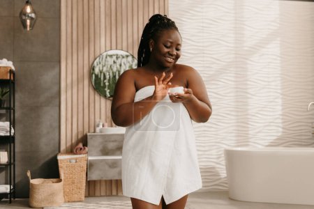 Téléchargez les photos : Happy plus taille femme africaine recouvert de serviette contenant avec crème visage dans la salle de bain - en image libre de droit