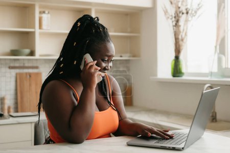 Foto de Alegre más tamaño mujer africana hablando en el teléfono móvil y el uso de la computadora portátil mientras trabaja en casa - Imagen libre de derechos