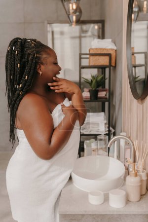 Foto de Confiado más tamaño mujer africana tocando la cara e irradiando amor propio mientras se mira al espejo en el baño - Imagen libre de derechos