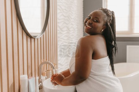 Foto de Alegre más tamaño mujer africana cubierta de toalla lavándose las manos y sonriendo en el baño doméstico - Imagen libre de derechos