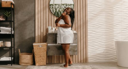 Foto de Hermosa mujer africana de tamaño grande cubierta con la cara de limpieza de toallas con almohadilla de algodón en el baño doméstico - Imagen libre de derechos