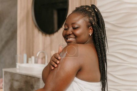 Téléchargez les photos : Belle femme africaine plus la taille couverte de serviette appliquer crème hydratante sur l'épaule dans la salle de bain - en image libre de droit