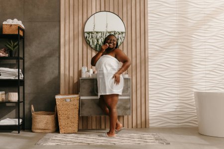 Foto de Feliz más tamaño mujer africana cubierta de toalla disfrutando de tratamiento de belleza en el baño doméstico - Imagen libre de derechos
