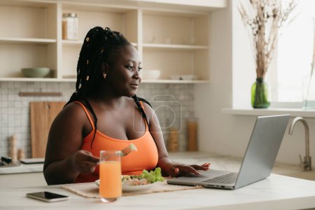 Téléchargez les photos : Belle femme africaine taille plus travaillant sur ordinateur portable tout en profitant d'une alimentation saine pour le déjeuner à la maison - en image libre de droit