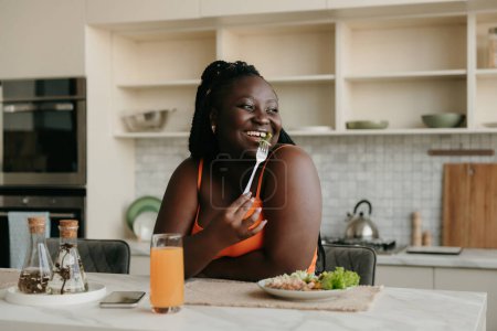 Foto de Hermosa mujer africana con curvas disfrutando de comida saludable para el almuerzo en la cocina doméstica - Imagen libre de derechos
