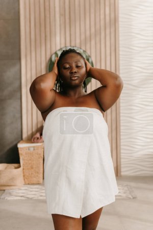 Téléchargez les photos : Belle femme africaine plus la taille couverte de serviette ajuster les cheveux tout en se tenant debout dans la salle de bain domestique - en image libre de droit
