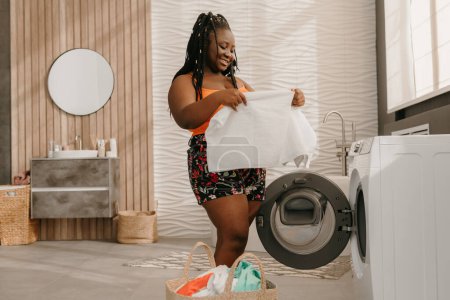 Photo for Happy plus size African woman loading clothes into washing machine while standing in domestic bathroom - Royalty Free Image