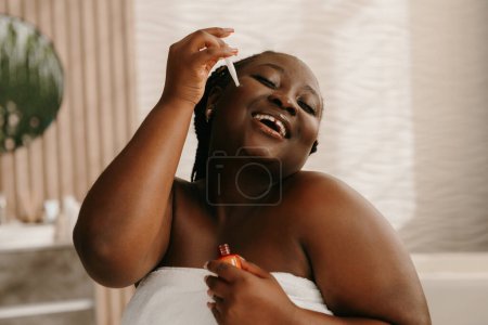 Téléchargez les photos : Heureuse femme africaine dodue recouverte de serviette appliquer sérum cosmétique sur le visage dans la salle de bain - en image libre de droit