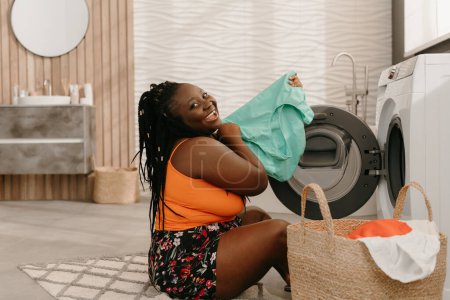 Photo for Happy plus size African woman holding fresh and clean clothes near the washing machine in bathroom - Royalty Free Image