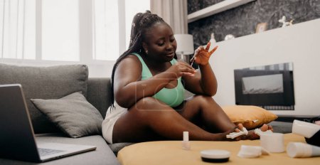 Photo for Beautiful plus size African woman painting toenails while sitting on the comfortable couch at home - Royalty Free Image