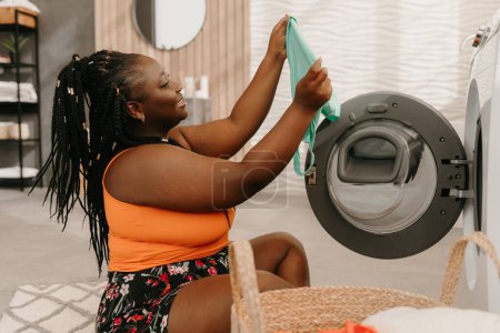 Photo for Happy plus size African woman examining fresh and clean clothes near the washing machine in bathroom - Royalty Free Image