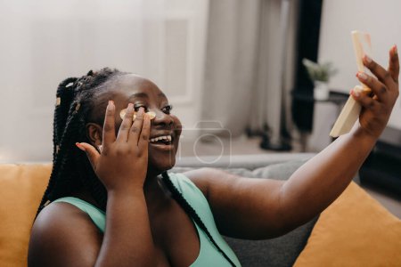 Téléchargez les photos : Happy plus taille femme africaine appliquant des patchs pour les yeux et regardant le miroir tout en se relaxant à la maison - en image libre de droit