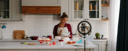 Téléchargez les photos : Confiant jeune femme préparant crème pour gâteau fait maison et enregistrement vidéo pour blog alimentaire - en image libre de droit