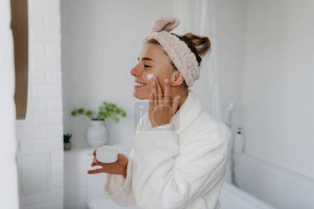 Téléchargez les photos : Joyeux jeune femme en peignoir appliquant crème visage tout en se tenant dans la salle de bain - en image libre de droit