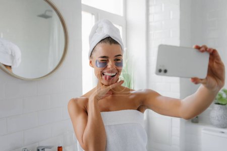 Téléchargez les photos : Jeune femme ludique couverte de serviette et avec des patchs sur les yeux faire selfie par téléphone intelligent dans la salle de bain - en image libre de droit