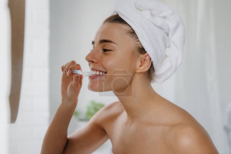 Foto de Mujer joven feliz con toalla sobre la cabeza cepillarse los dientes en el baño - Imagen libre de derechos