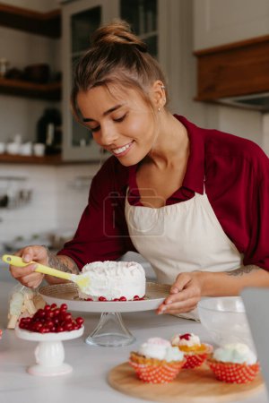 Foto de Feliz joven pastelera aplicando crema batida mientras decora la torta en la cocina doméstica - Imagen libre de derechos