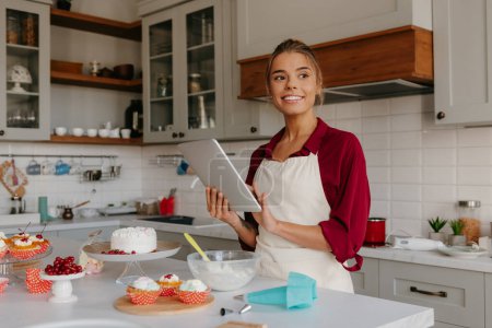 Foto de Hermosa joven pastelera usando tableta digital mientras prepara pasteles y magdalenas en la cocina - Imagen libre de derechos