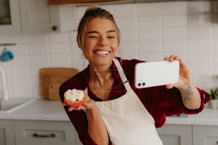 Photo for Joyful female baker using smart phone while making selfie with freshly made muffin on the kitchen - Royalty Free Image