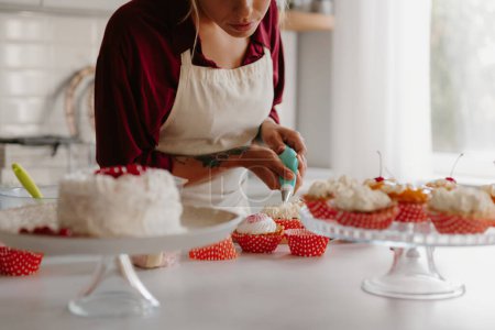 Photo for Close-up of female confectioner squeezing whipped cream while decorating muffins on the kitchen - Royalty Free Image