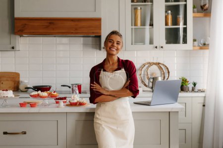 Photo for Happy female baker looking confident while standing near the kitchen island with variety of sweet food - Royalty Free Image