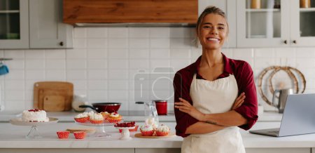 Photo for Beautiful female baker looking happy while standing near the kitchen island with variety of sweet food - Royalty Free Image