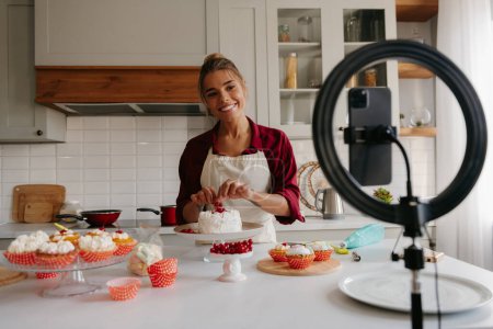 Téléchargez les photos : Confiseur féminin heureux décorant gâteau sur la cuisine tout en filmant le processus sur téléphone intelligent - en image libre de droit