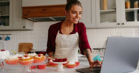 Foto de Hermosa pastelera femenina usando el ordenador portátil mientras hace pasteles y magdalenas en la cocina - Imagen libre de derechos