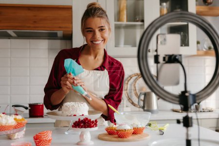 Téléchargez les photos : Boulanger féminin confiant appliquant de la crème fouettée sur le gâteau tout en streaming en ligne de la cuisine - en image libre de droit