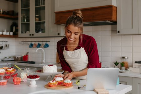 Foto de Hermosa hembra panadero merchandising muffins y sonriendo mientras que de pie en la cocina doméstica - Imagen libre de derechos