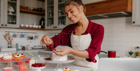 Foto de Pastel de confitería femenina feliz decoración con bayas, mientras que de pie en la cocina doméstica - Imagen libre de derechos