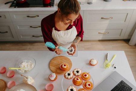 Foto de Vista superior del confitero femenino aplicando crema batida sobre los pasteles mientras está de pie en la cocina - Imagen libre de derechos