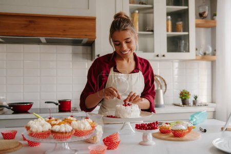 Foto de Hermosa pastelera femenina que decora el pastel con bayas mientras está de pie en la cocina doméstica - Imagen libre de derechos