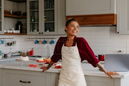 Photo for Beautiful female baker looking happy while leaning at the kitchen island with variety of sweet food - Royalty Free Image