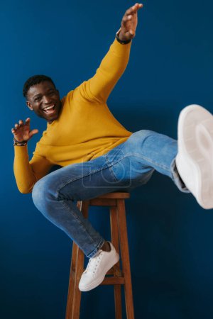 Téléchargez les photos : Jeune homme africain ludique donnant des coups de pied à la jambe et gesticulant assis sur la chaise contre le mur bleu - en image libre de droit