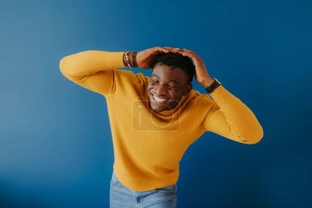 Photo for Joyful young African man in casual wear holding head in hands and smiling while standing on blue background - Royalty Free Image