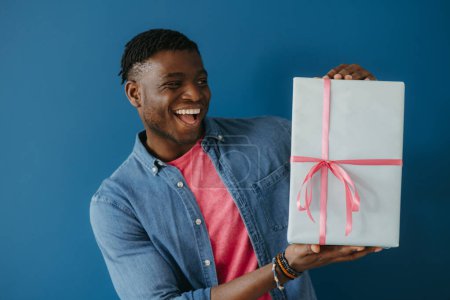 Téléchargez les photos : Heureux jeune homme africain en tenue décontractée tenant boîte cadeau et souriant sur fond bleu - en image libre de droit