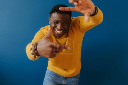 Téléchargez les photos : Jeune homme africain ludique en tenue décontractée se concentrer sur son visage tout en se tenant debout sur fond bleu - en image libre de droit