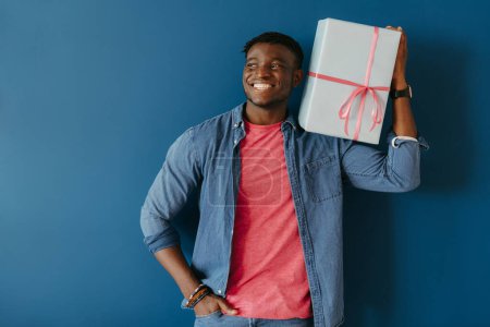 Photo for Handsome young African man in casual wear carrying gift box on shoulder and smiling on blue background - Royalty Free Image